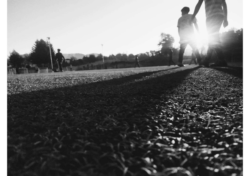 Siluetas de personas caminando sobre un campo de fútbol al atardecer, con sombras largas y luz brillante en el fondo.