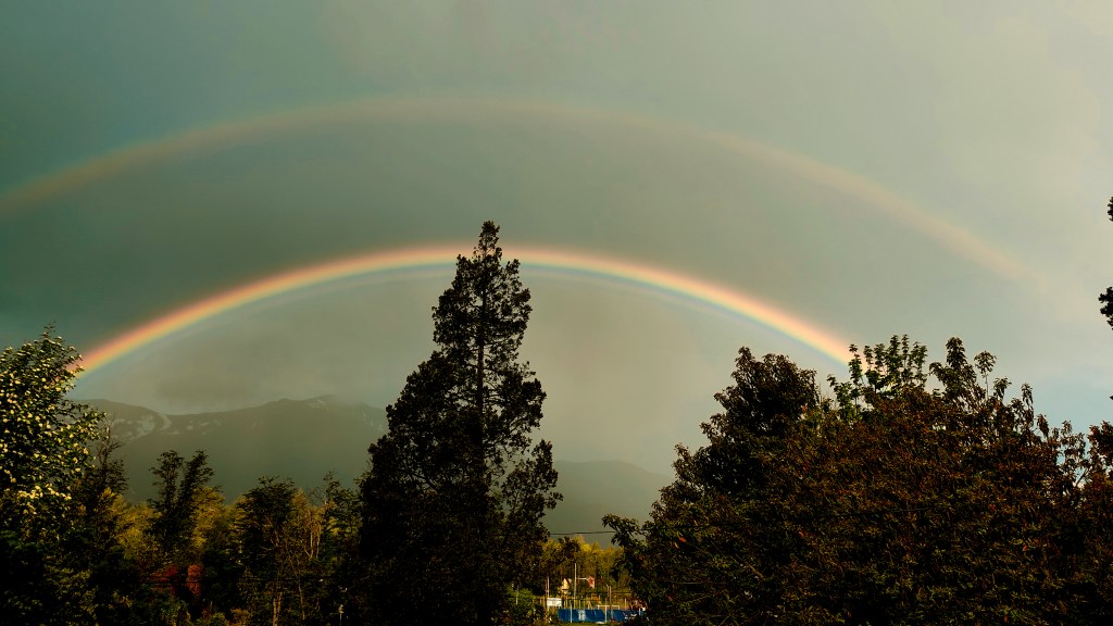 Arco iris doble sobre un paisaje montañoso, con árboles en primer plano.