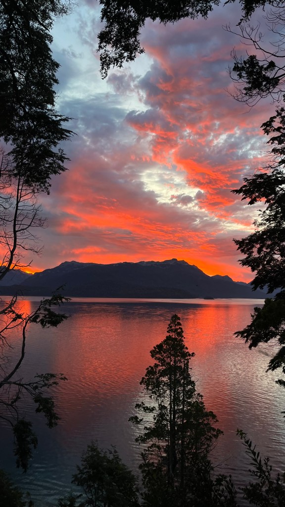 Atardecer sobre un lago rodeado de montañas y árboles, con cielos llenos de nubes rosas y naranjas.